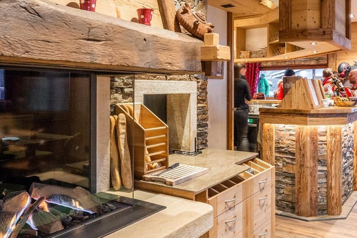 A cozy, rustic chalet-style interior featuring a modern glass fireplace, a bread display, and a stone and wood counter. People are in the background.