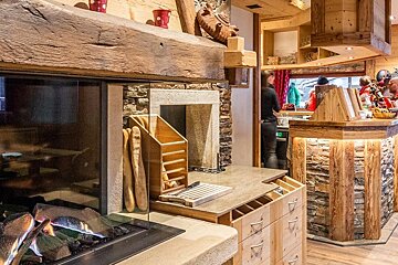 A cozy, rustic chalet-style interior featuring a modern glass fireplace, a bread display, and a stone and wood counter. People are in the background.