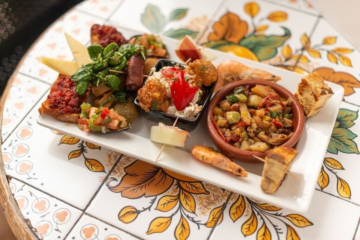 A vibrant platter of assorted tapas including shrimp, bruschetta, and various small dishes, served on a white tray over a decorative tiled table.