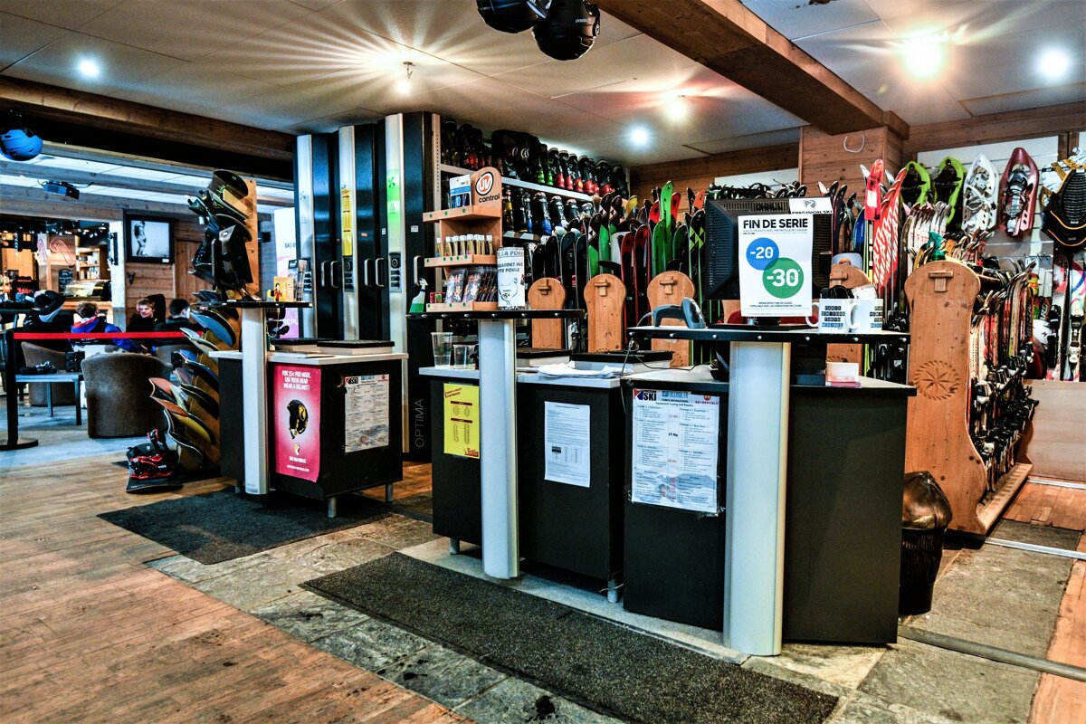 A bustling ski and snowboard rental shop interior, featuring multiple service counters, extensive racks of skis, snowboards, boots, and helmets.