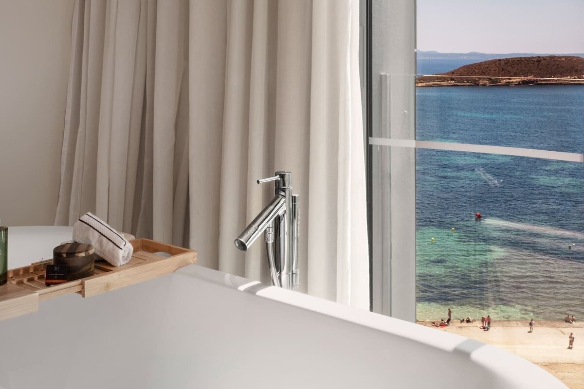 A sleek bathtub with bath accessories overlooks a stunning ocean and sandy beach through a large window, framed by light curtains.