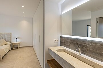 A modern bedroom features white built-in wardrobes alongside a sleek concrete vanity with a chrome faucet, illuminated mirror, and a tiled backsplash.