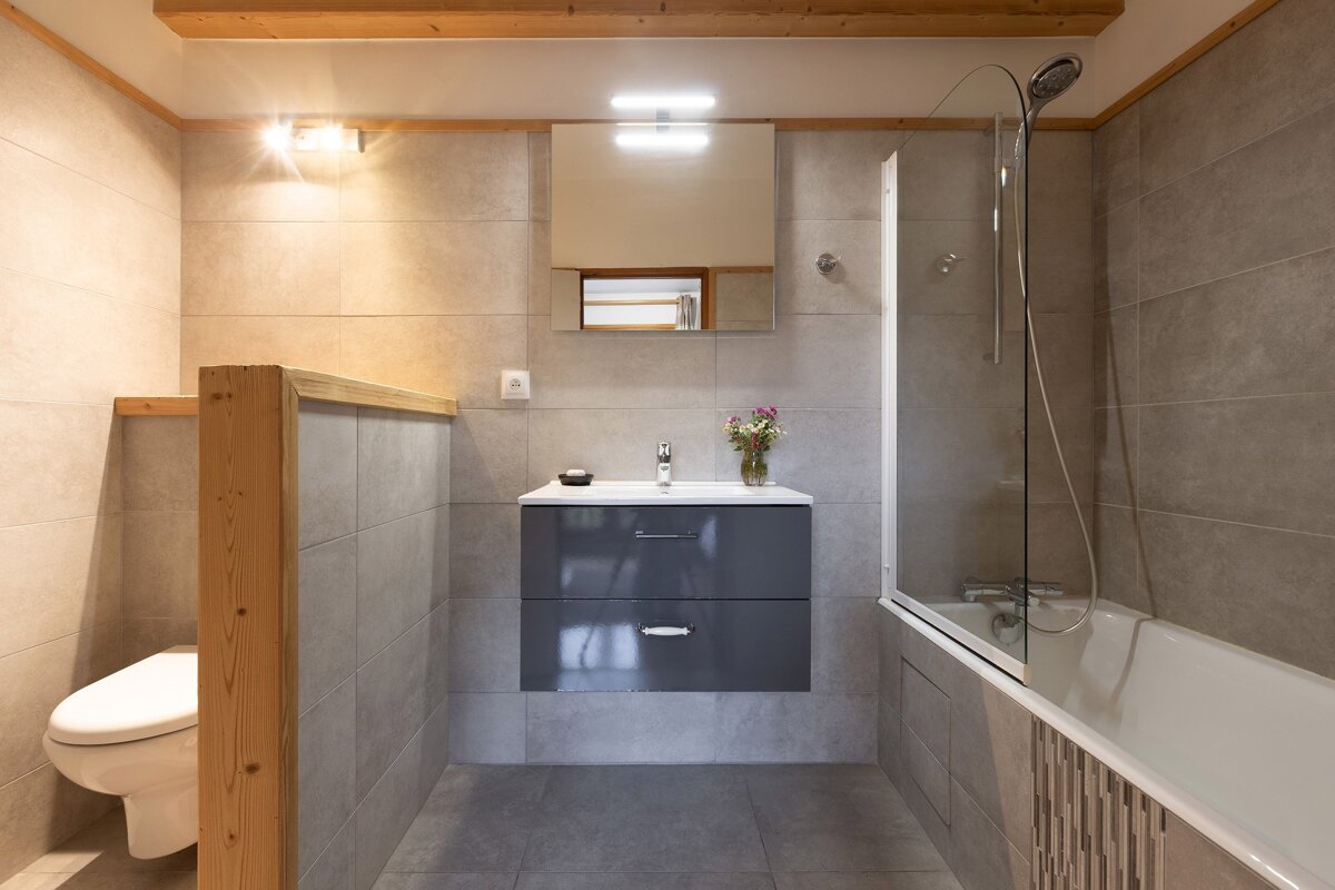 A modern bathroom features gray tiled walls and floor, a toilet partially hidden by a wooden half-wall, a dark vanity with sink, and a bathtub with a glass shower screen. Wooden beams add warmth.