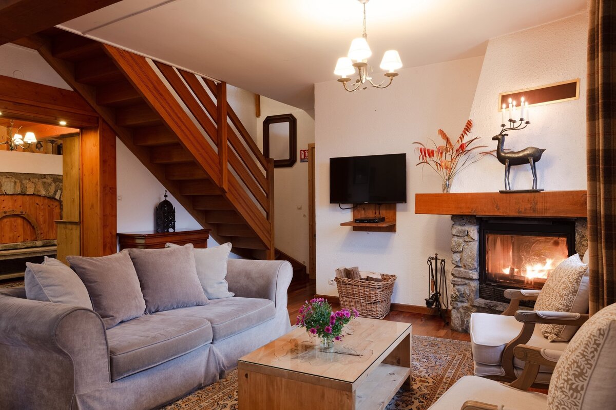A cozy living room with a lit stone fireplace, a grey sofa, wooden staircase, TV, and coffee table with flowers.