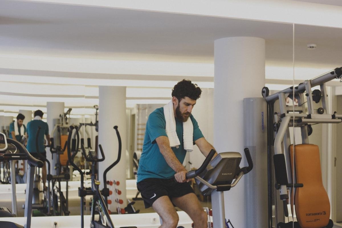 A bearded man with a towel around his neck cycles on a stationary bike in a bright gym with mirrors reflecting other equipment.