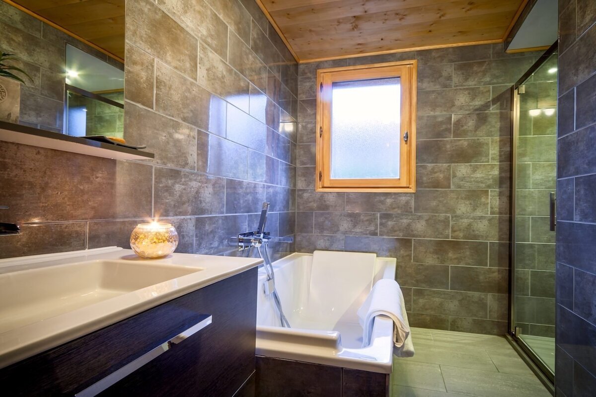 A rustic bathroom features dark tiled walls, a wooden ceiling, a sink with a lit candle, a bathtub, a window, and a shower.