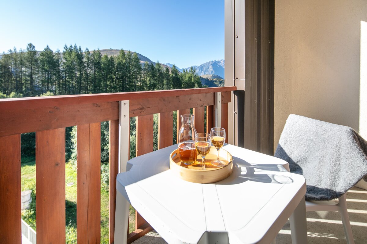 A sunny balcony with drinks on a table and a chair, offering a scenic view of a pine forest, a lush mountain valley, and distant peaks.