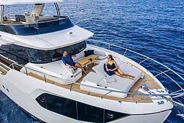 A man and woman relax on the foredeck of a modern white yacht, enjoying the calm blue sea.
