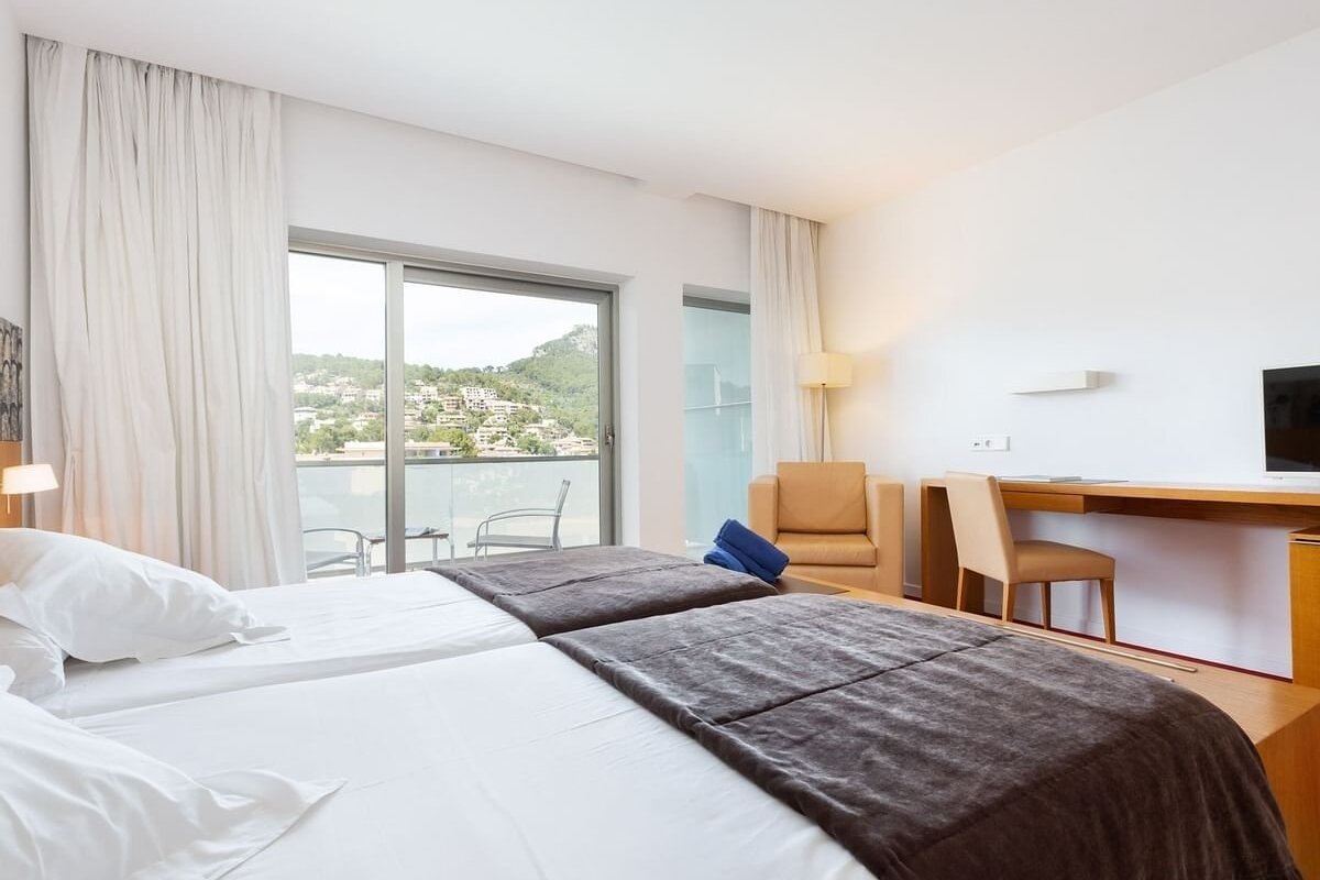Bright modern hotel room with twin beds, a desk, and a balcony overlooking a hillside town.
