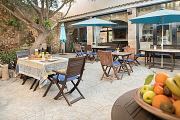 An inviting outdoor restaurant patio with wooden tables, blue umbrellas, a breakfast setting, and a large tree next to a building.