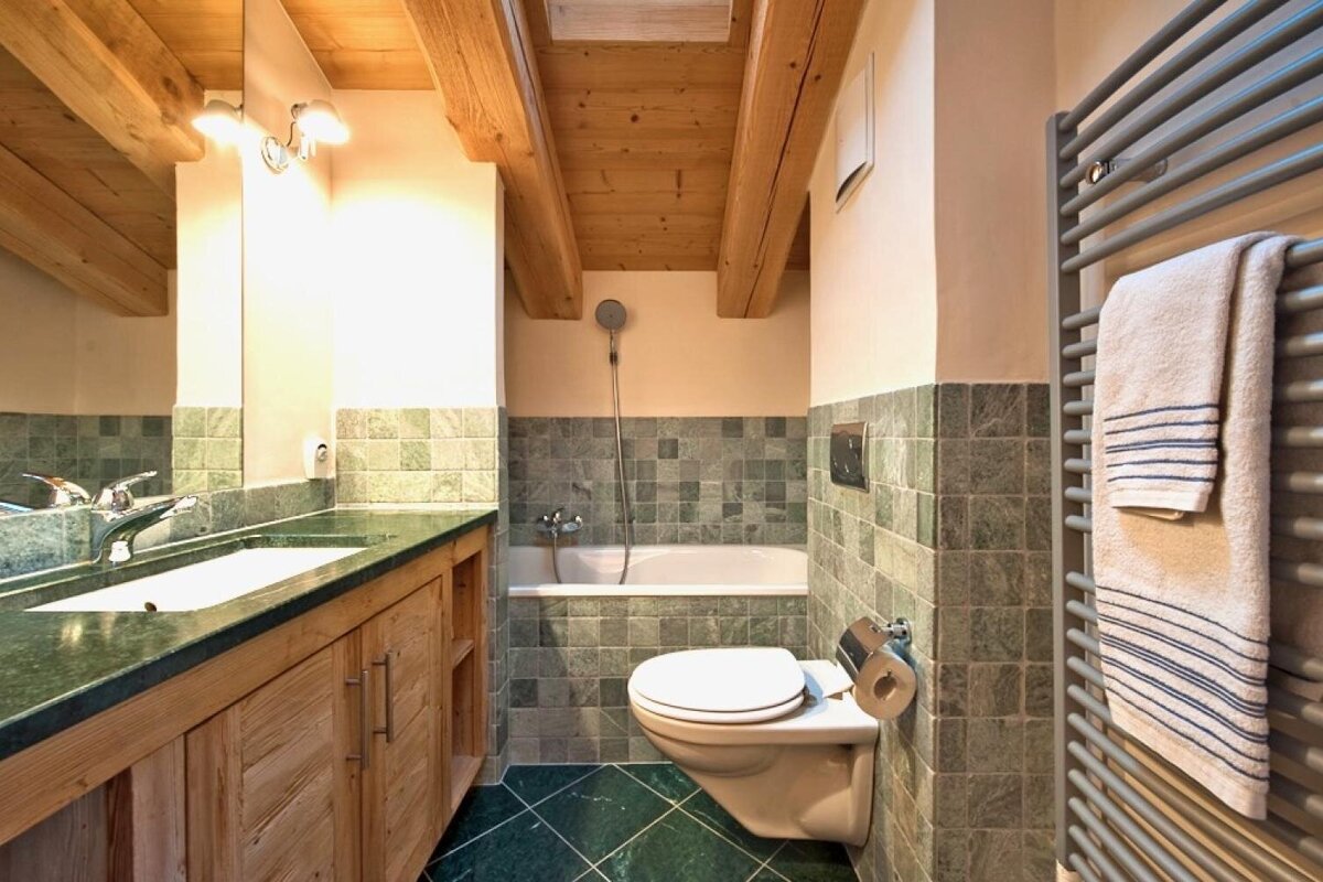 A cozy, rustic bathroom featuring exposed wooden beams, green tiled walls and floor, a bathtub, floating toilet, and wooden vanity.