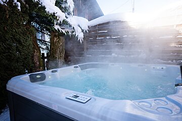 A steaming hot tub in a snowy outdoor setting. Snow-covered trees and a wooden fence are visible, with sunlight filtering through the mist.