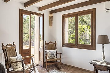 A bright room with white walls, wooden beams, a glass door open to trees, a window, and two traditional wooden chairs on a tiled floor.