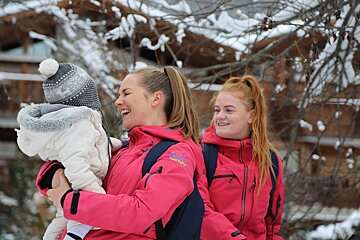 Two women in pink jackets, one holding a baby in a snowsuit and hat, all smiling joyfully in a snowy winter landscape.
