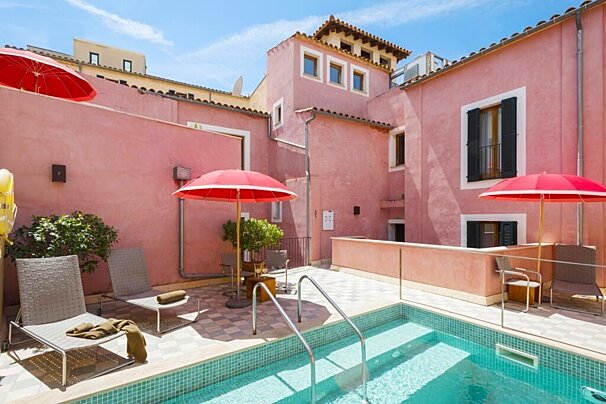 A vibrant pink building frames a sunny rooftop terrace with a small, tiled swimming pool, lounge chairs, and bright red umbrellas.