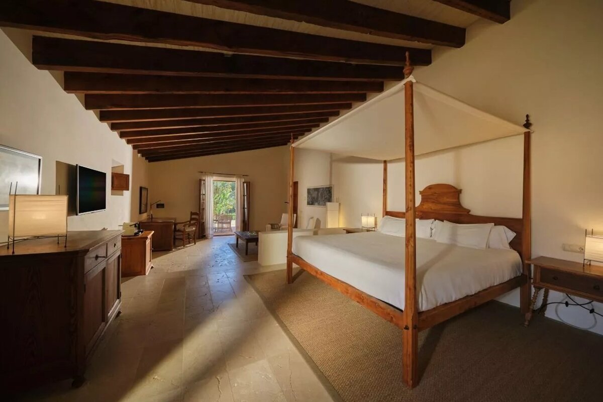 A large, rustic bedroom with a wooden four-poster bed, exposed ceiling beams, and traditional decor. A door opens to an outdoor space.