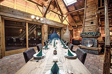 Rustic lodge dining room with a long table set for guests under a high beamed ceiling, featuring a cozy, decorated fireplace.