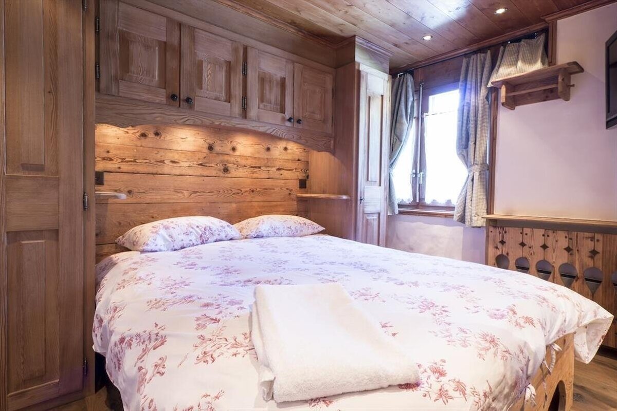 A cozy, rustic wooden bedroom featuring a double bed with floral bedding, built-in wooden cabinets, and a window with curtains.