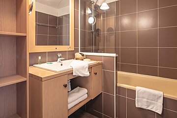 A brown-tiled bathroom featuring a wooden vanity with a sink, mirror, and shelves. A shower-bath combo with a glass screen and towels is visible.