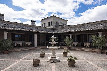 A charming L-shaped courtyard features a tiered fountain, potted plants, and covered patios with dining tables under a cloudy sky.