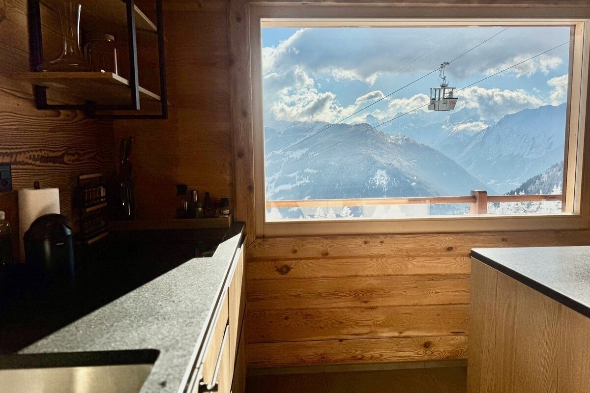 Cozy wooden room overlooking snowy mountains with a ski lift, under a bright blue sky with clouds.