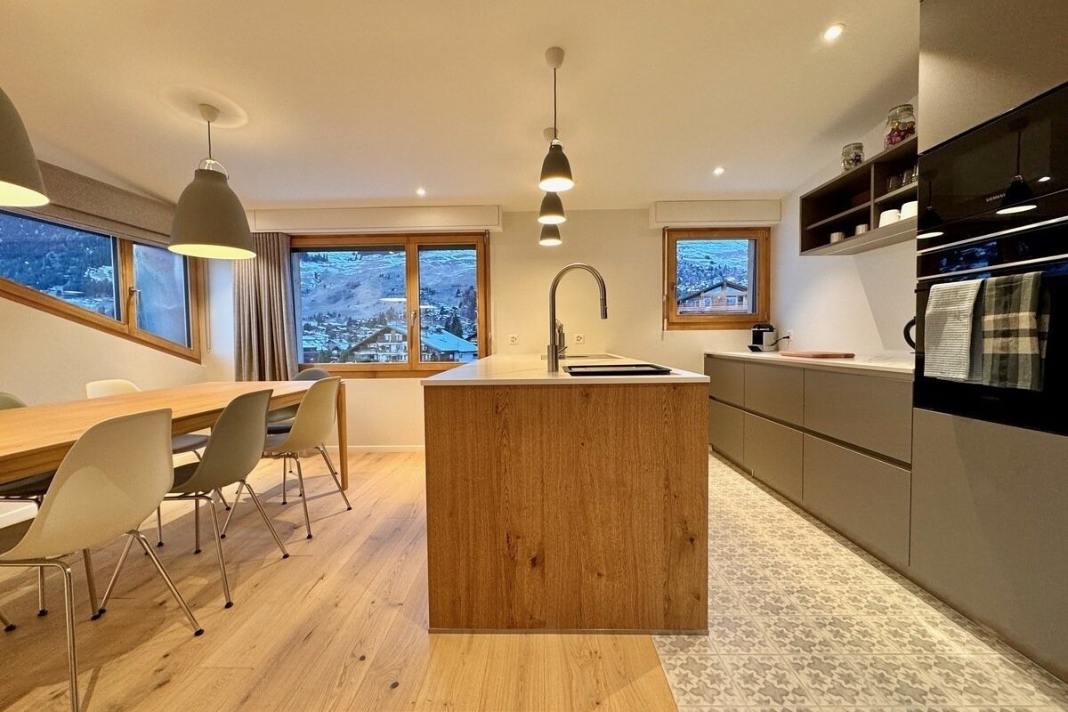 Chic kitchen and dining area with a wood island, sleek cabinets, and a dining table. Large windows overlook a snowy mountain village.