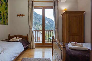 A cozy twin bedroom with wooden beds and furniture, featuring a balcony door leading to a stunning mountain view. White bedding and warm wood create a rustic ambiance.