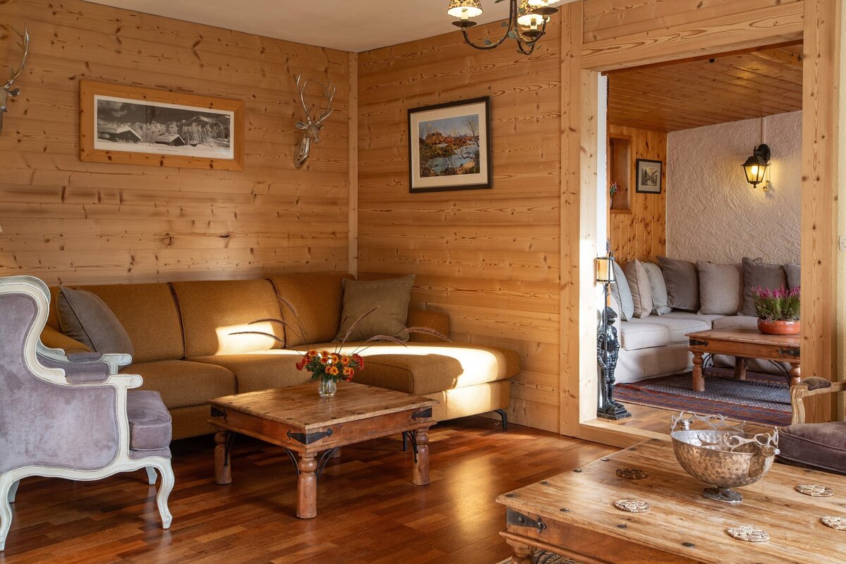 A rustic, wood-paneled chalet living room with a brown sectional, wooden tables, and antler decor. Warm light illuminates the cozy space, revealing another seating area.
