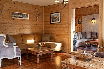 A rustic, wood-paneled chalet living room with a brown sectional, wooden tables, and antler decor. Warm light illuminates the cozy space, revealing another seating area.