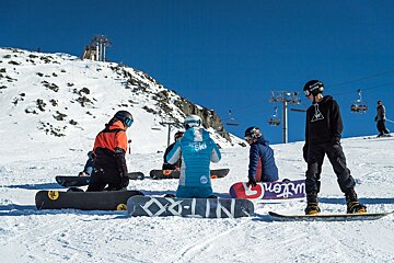 Snowboarders on a sunny, snowy mountain with a clear blue sky. Several are sitting or kneeling on their boards, while one stands, with ski lifts in the background.