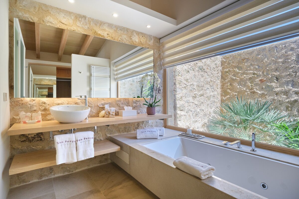 A chic stone-walled bathroom with a vessel sink, large mirror, and a modern bathtub by a window showing green plants. Features neutral tones.