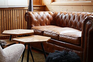 A cozy room with a tufted brown leather sofa, two hexagonal wooden tables with herringbone tops, and a fluffy white chair. Wooden paneling adds warmth.