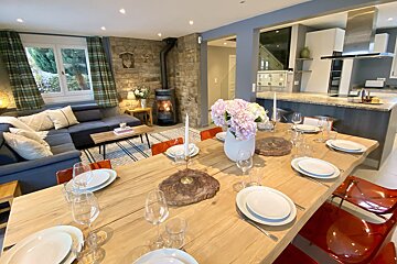 A cozy, open-plan home featuring a large set dining table, a living room with a lit wood stove, and a modern kitchen.