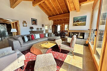 Bright, cozy living room featuring vaulted wooden ceilings, a large sectional sofa, stone fireplace, unique coffee table, and a dining area.