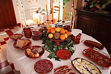 A bountiful table spread with a variety of foods like cold cuts, cheese, bread, and fresh fruit in earthenware dishes, adorned with candles and ivy.