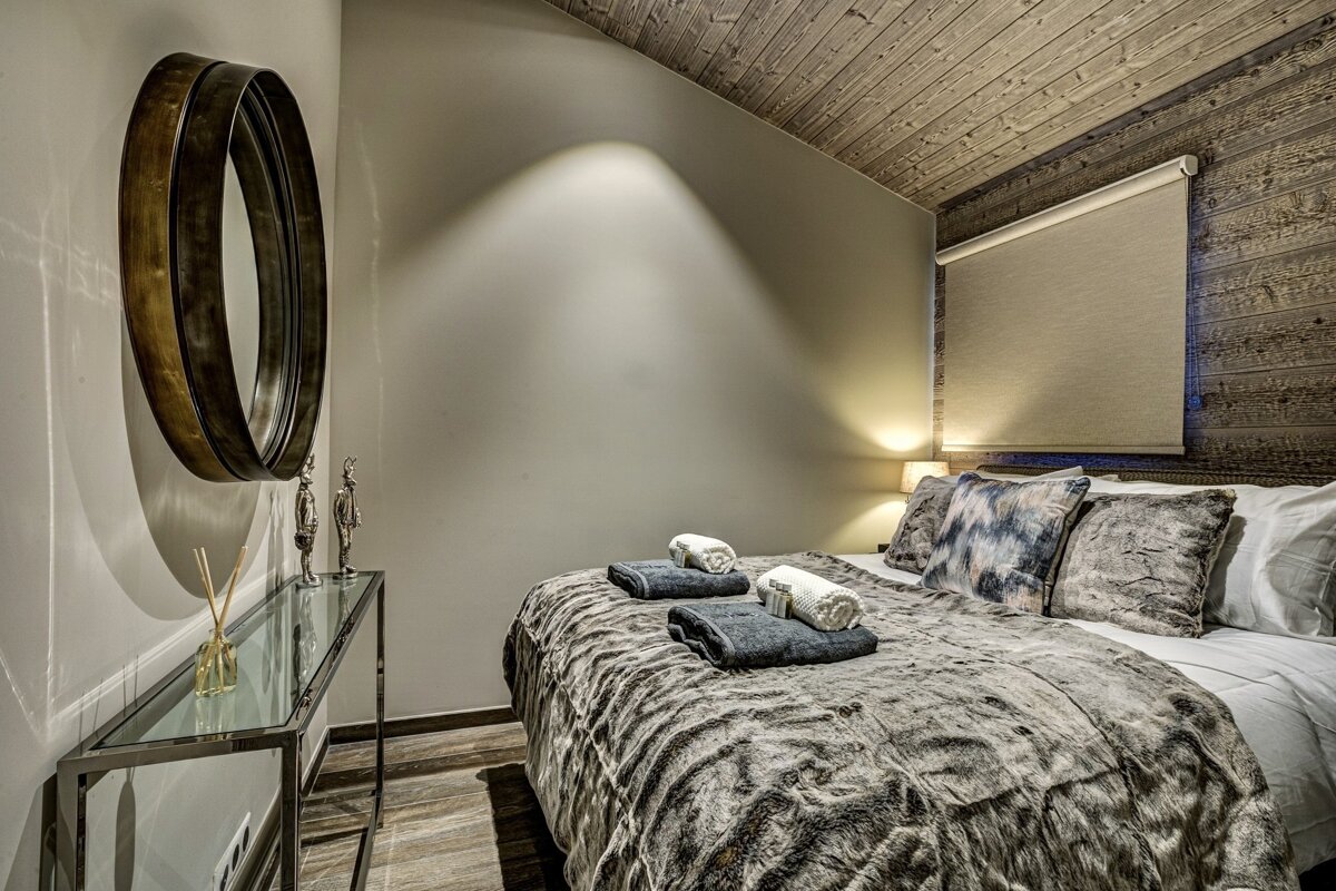 A cozy, rustic-chic bedroom featuring a faux fur bed, wood-paneled ceiling and wall, a large round mirror, and soft ambient lighting.
