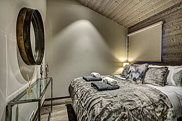 A cozy, rustic-chic bedroom featuring a faux fur bed, wood-paneled ceiling and wall, a large round mirror, and soft ambient lighting.