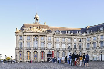 A grand, ornate building with a cupola stands under a clear sky. A long line of people waits in a cobblestone square in front of it.