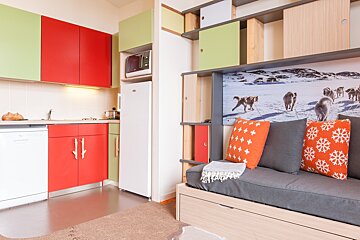 Cozy studio interior with a red and green kitchen next to a grey daybed featuring bright orange pillows and dog sled wall art.