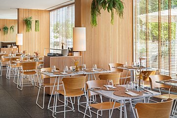 A modern restaurant interior with light wood walls, hanging plants, marble-top tables, and white chairs, featuring large windows with blinds.