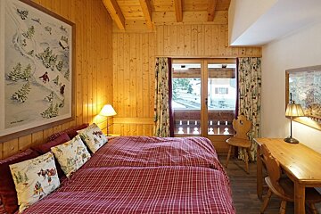 Cozy, rustic bedroom with wood paneling, red plaid bed, ski-themed art, a desk, and a balcony door revealing an outdoor view.