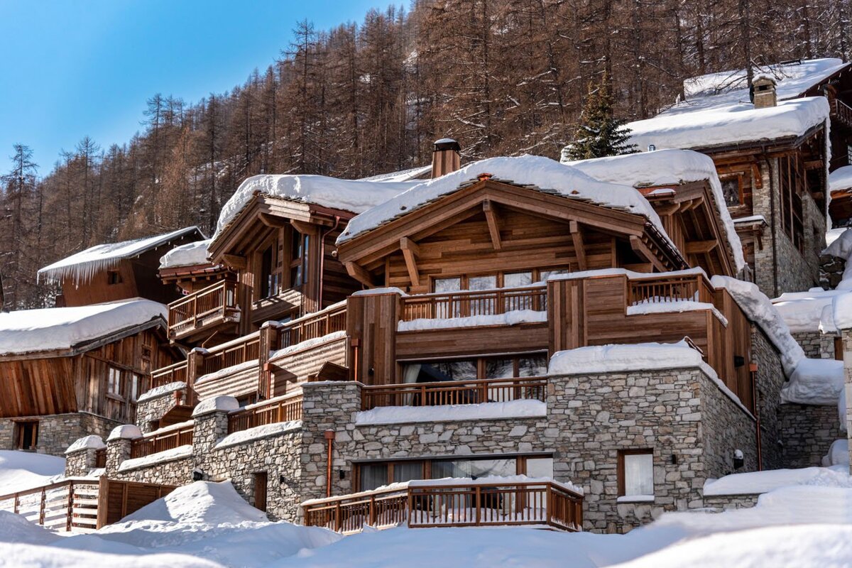 Rustic wooden and stone chalets, heavily snow-covered, built into a sunny, forested mountainside under a clear blue sky.