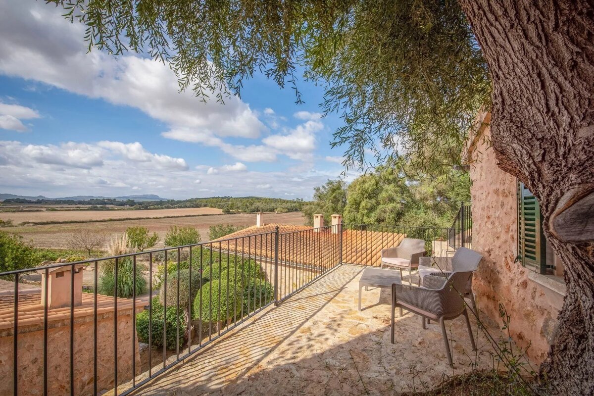 A sunny, rustic stone balcony with outdoor furniture and a railing, overlooking vast fields, trees, and hills under a partly cloudy sky. An old tree frames the view.
