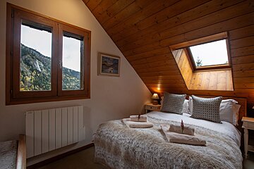 A warm, rustic attic bedroom features a sloped wooden ceiling, a bed with a furry throw, and windows offering a scenic mountain waterfall view.