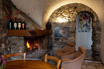 A cozy, rustic room features a roaring stone fireplace, wine bottles on the mantel, comfy armchairs, and a wooden table. Arched stone walls create an inviting ambiance.