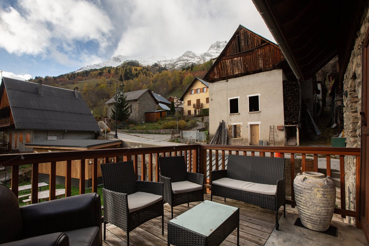A cozy balcony with wicker furniture overlooks an alpine village and colorful autumn trees, backed by snow-capped mountains under a partly cloudy sky.