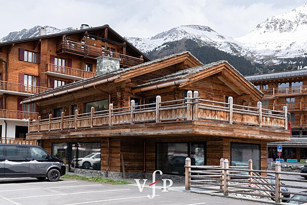 Rustic wooden chalet-style buildings with balconies against a backdrop of snowy mountains. A black van is parked in the foreground.