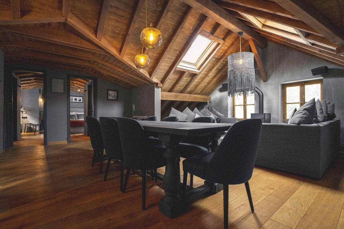 A spacious, rustic-modern attic apartment features exposed wooden beams, hardwood floors, a dining area, and a cozy living space with skylights and stylish lighting.