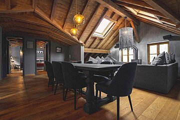 A spacious, rustic-modern attic apartment features exposed wooden beams, hardwood floors, a dining area, and a cozy living space with skylights and stylish lighting.