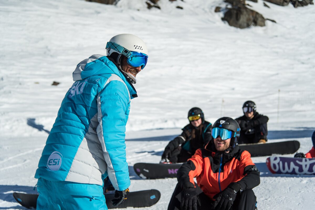 A person wearing a ski jacket talks to a group of snowboarders
